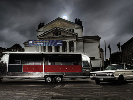 Vintage Anhänger und Oldtimer vor einem historischen Gebäude unter dunklem, wolkigem Himmel.
