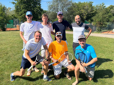 Sieben Männer mit Tennisschlägern posieren lachend auf einer Wiese vor Tennisplätzen an einem sonnigen Tag.
