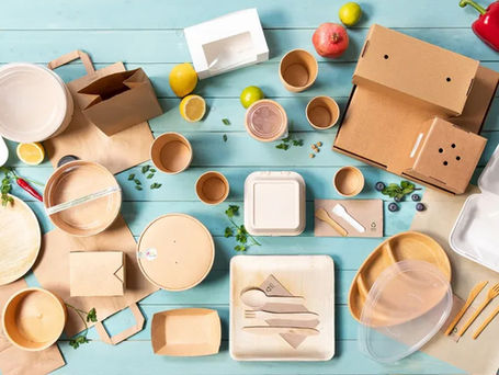 Umweltfreundliche Einweggeschirr auf hellblauem Tisch, umgeben von frischen Früchten und Gemüse. Pappbecher, Schalen und Recyclingkartons.