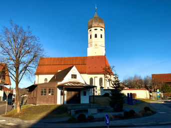 Dorfplatz von Erling mit der historischen Dorfschmiede und der Kirche St. Valentin bei blauem Himmel.
