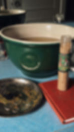 A photo taken by Austin Fuller of Bane X Bramble of amoney-drawing bath ritual with Money Drawing Wash Crystals sitting on top of a red magical book. The bath is in a green and white basin and sitting on top of a blue counter with incense burning on a silver plate.