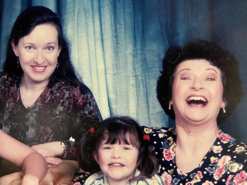 Studio photo showing a white woman smiling into the camera with long dark hair in her early 30s, a little white girl of about 2 and a half with dark hair in bunches, also smiling at the camera, and an older woman around 60 with short black hair, laughing into the camera.