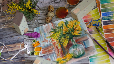 A colorful workspace features a watercolor painting of yellow flowers in a vase, surrounded by paint swatches, brushes, sunglasses, and small figurines.