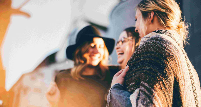 Three women stand together, smiling and laughing in front of a building, part of a lively laughter ESL lesson plan.