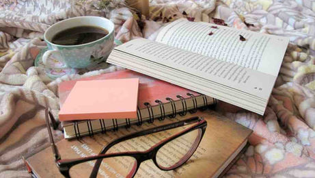 A book, reading glasses, and a cup of coffee are arranged on a neatly made bed.