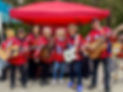 Mechanics of Music adult guitar students in matching red jerseys performing at a community event under a red tent.