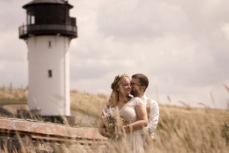 Bräutigam hat seine Braut im Arm und im Hintergrund ist ein kleiner Leuchtturm zu sehen