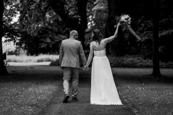 Brautpaar läuft Hand in Hand im Park entlang und die Braut hält ihren Strauß fröhlich hoch.