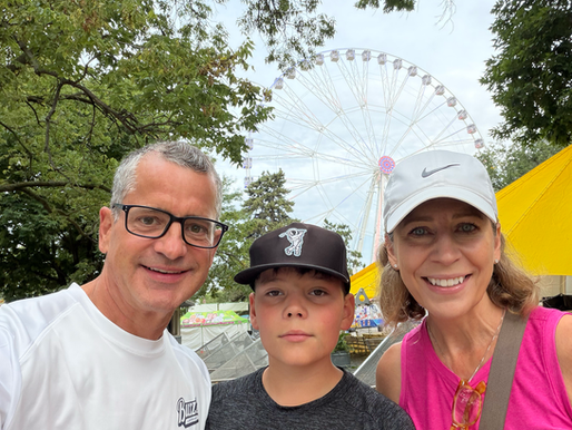 Family selfie at a fair, smiling before a giant Ferris wheel, framed by trees and yellow tents.