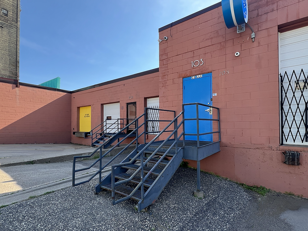 Terracotta, lay-colored brick industrial building with blue door and metal stairs, a yellow dock door, and unit numbers 103 and 104, under a clear sky.