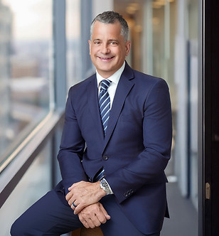 Smiling man in a blue suit with striped tie. Sits on a stool in front of a blurred office window background.