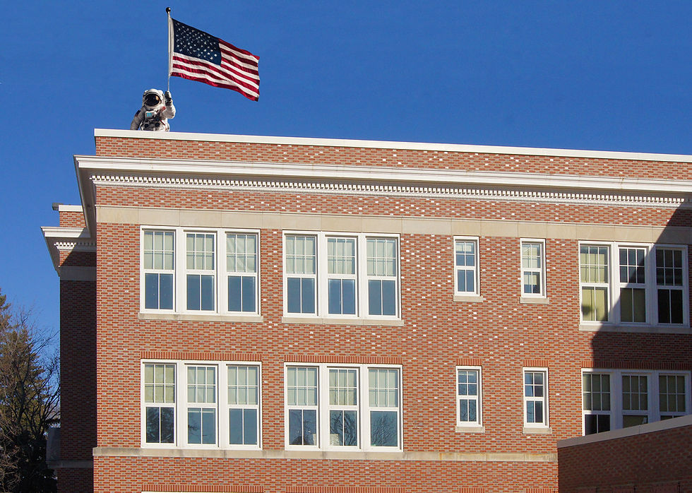 Astronaut flying a US flag on the roof of a brick school with windows.