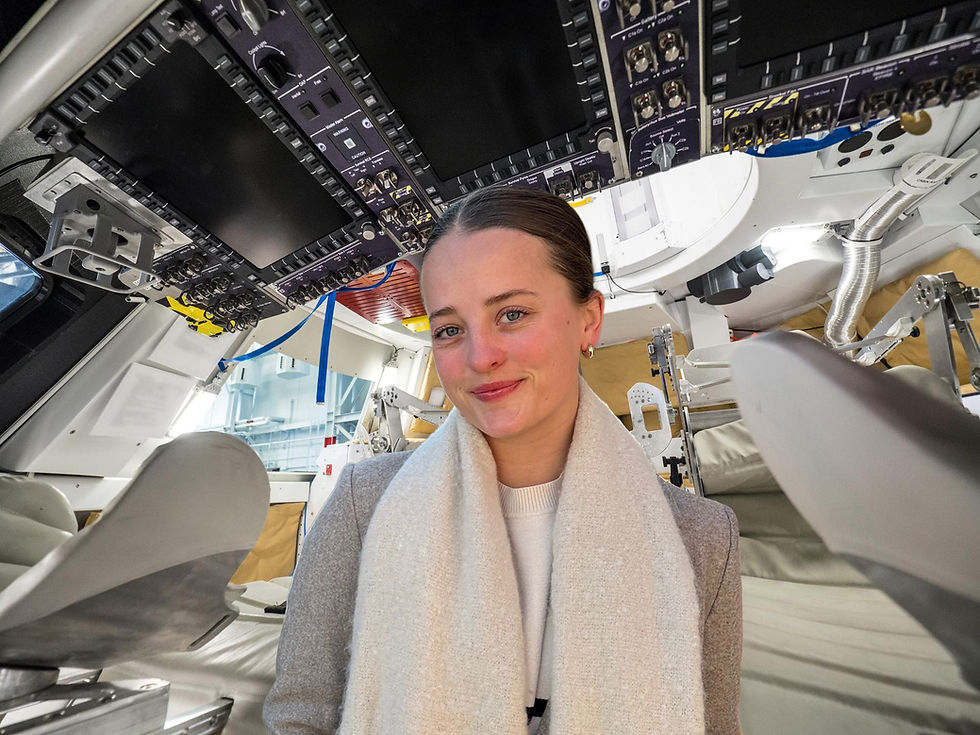 Halie smiling inside a space capsule, surrounded by control panels and seats. The background is metallic with blue and beige accents.