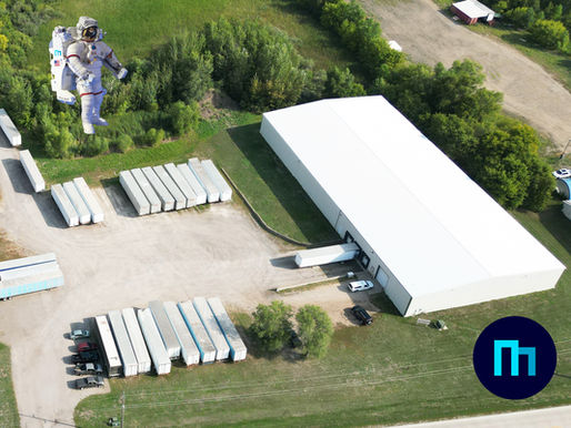 Aerial view of a warehouse and lot with trailers and vehicles. An astronaut floats above grassy terrain. Blue logo in the corner. Peaceful, yet productive setting.