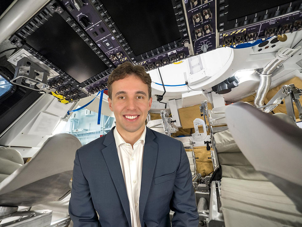 A smiling man in a suit is inside a spacecraft cockpit with seats and control panels. Background has tubes and machinery.