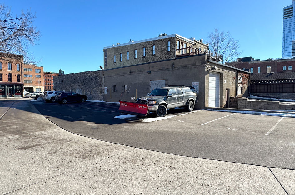 4. The rear exterior of a concrete block building features a white roll-up garage door and a small service entrance. A black pickup truck is parked in the adjacent asphalt lot, which has visible parking stripes and some lingering patches of snow. Behind the building, a multi-story brick structure with wooden balconies is visible under a clear blue sky.