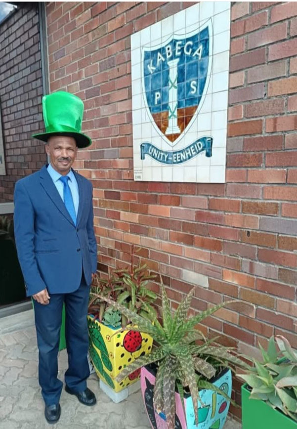 Man in a blue suit with a green hat stands by colorful planters. A tiled sign reads "Kabega PS Unity-Eenheid" on a brick wall.