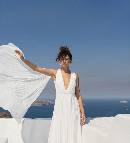 A side view of a model in a floor-length REPULO’S pleated wedding gown featuring a structured bodice and open-back detail. She is styled with long black opera gloves in a sunlit, rustic indoor setting with vintage wooden doors.