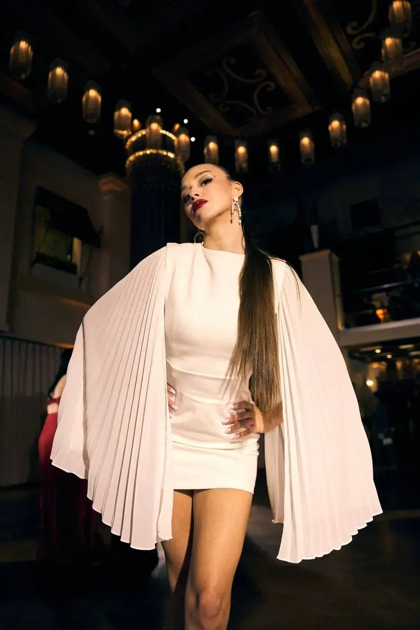 Модель демонструє білу коктейльну міні-сукню «Каталіна» з плісированими рукавами-крилами під час модного показу бренду REPULO'S у Торонто. Фото передає атмосферу вечірнього івенту з розкішними люстрами на задньому плані."
