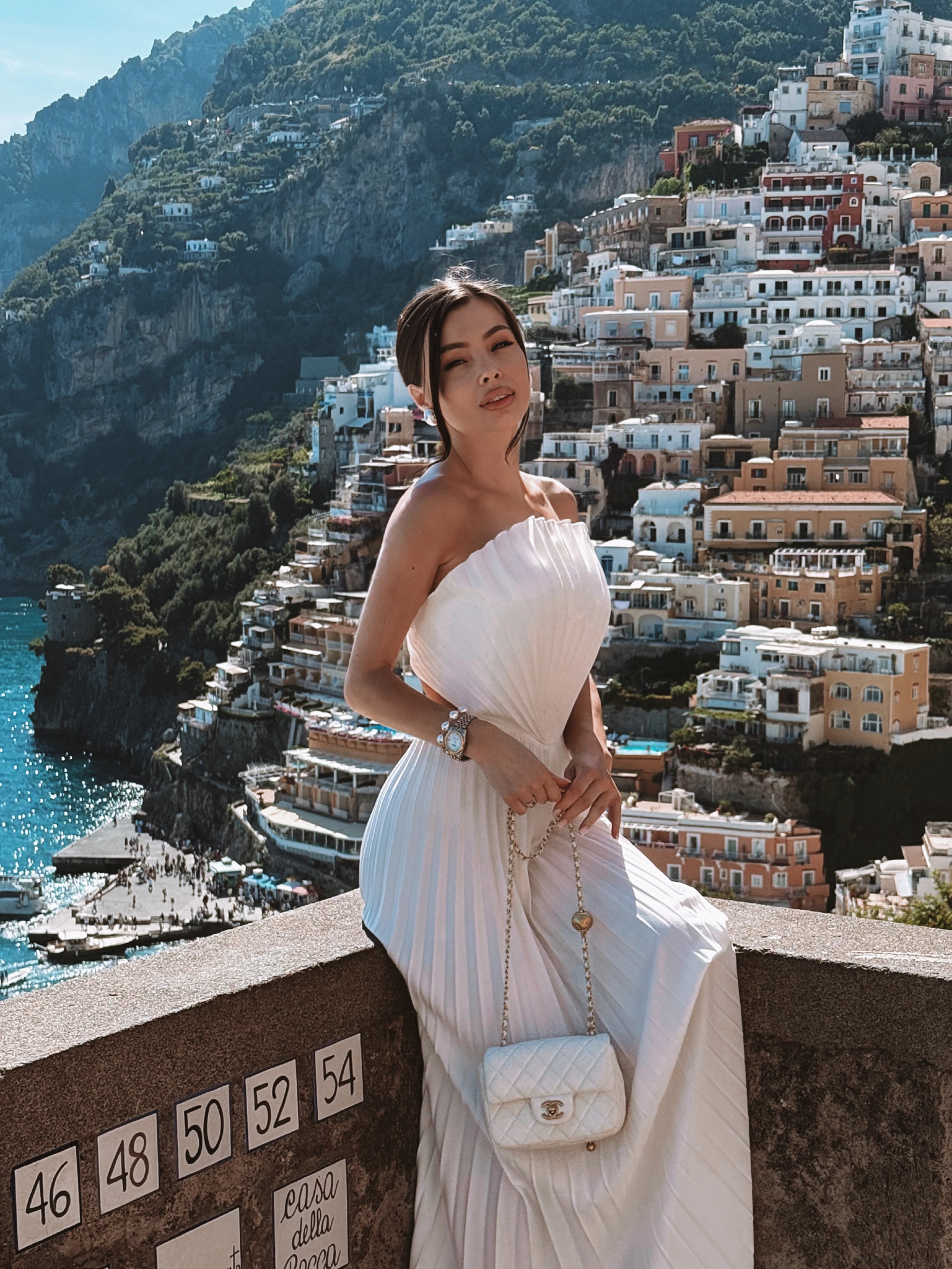 Back view of white pleated maxi dress by REPULO'S seated on Positano terrace overlooking turquoise Amalfi Coast water