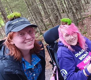 Two white disabled women (one standing, another in a wheelchair) smiling.