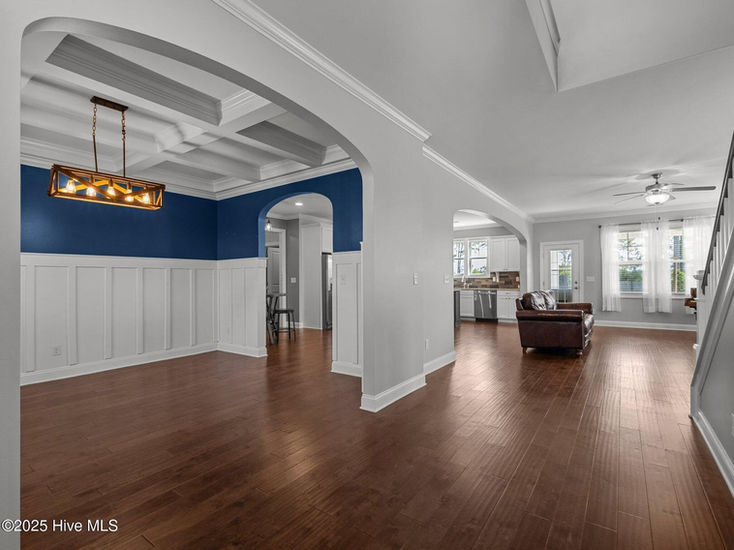 Spacious open-concept interior featuring rich hardwood floors, white wainscoting, a deep blue accent wall, coffered ceiling in the dining area, and natural light flowing into the adjacent living space.