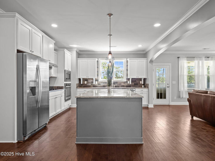 Modern kitchen featuring a large gray center island, white cabinetry, stainless steel appliances, tile backsplash, pendant lighting, and an open layout connecting to the living area.