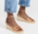Close-up of feet in tan wedge sandals with ankle straps and logo detail, worn with cuffed blue jeans on a white background. Walking Shoes for Las Vegas.