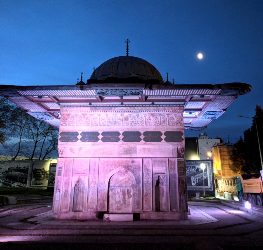 Ornate stone pavilion with intricate carvings, illuminated in pink and purple hues, under a twilight sky with a bright moon. Urban backdrop.