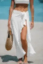 Woman in a white wrap skirt walks on a sunny beach, holding a woven tote, with blue water and sand behind her. Perfect for traveling wrinkle free beach wear.