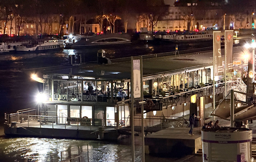 Night view of "Le Bistro Parisien" on a boat by a river. Lit interiors show diners inside. Reflective water, city lights in the background. Seine river dinner cruise. Top Things to do in Paris.