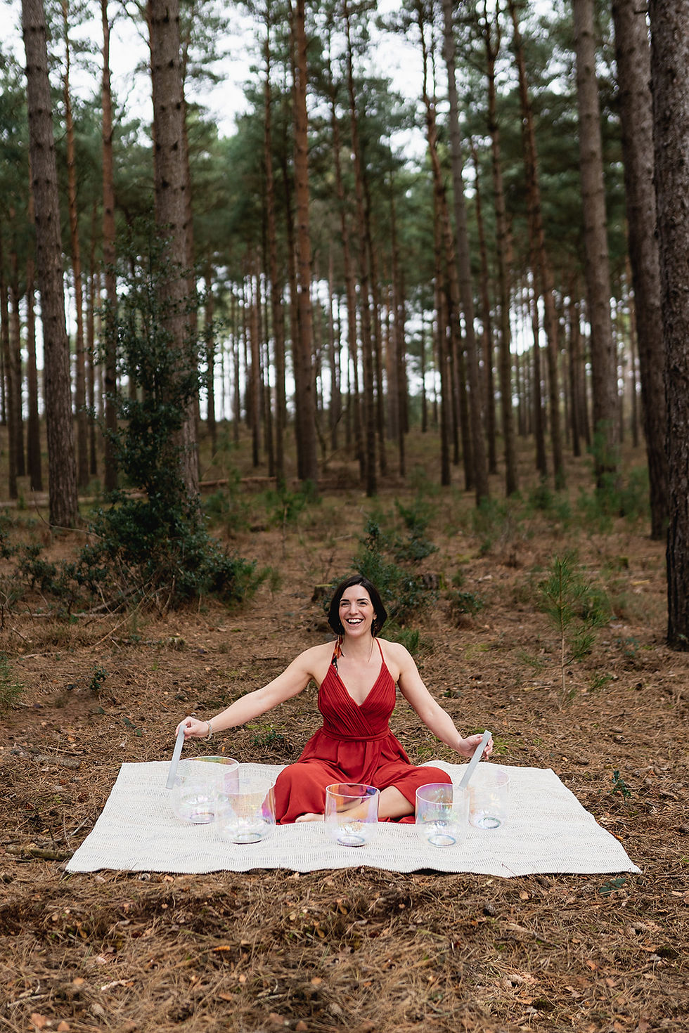 Kate in a red dress sits on a white blanket in a forest, playing crystal singing bowls.