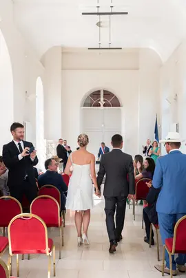 Photo d'un couple de mariés avançant dans l'allée d'honneur d'une lumineuse salle des mariages de la mairie.