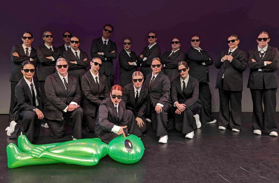 Un groupe de danseurs adultes sur scène, vêtus de costumes noirs assortis et portant des lunettes de soleil, s'agenouillent ensemble, tandis qu'un extraterrestre vert gonflable est allongé sur le sol devant eux.