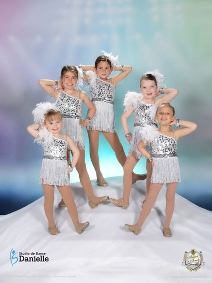 Un groupe de cinq jeunes danseurs de claquettes vêtus de robes blanches à plumes posant devant une toile de fond vibrante.