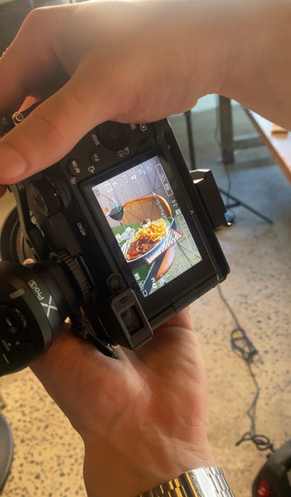 View through a camera screen showing a styled food shoot in progress, capturing a beautifully plated dish with studio lights and crew blurred in the background.