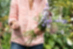 Person in a pink shirt holds a bouquet of purple and white flowers, tying stems with string, set in a lush green garden.