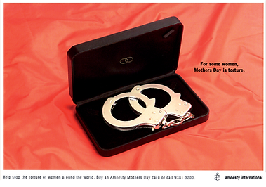 A black velvet box opens to reveal shiny handcuffs resting inside, against a vivid red background. The text reads, "For some women, Mothers Day is torture". This artwork by John Sander conveys a powerful message for the Amnesty International press campaign addressing women's rights.