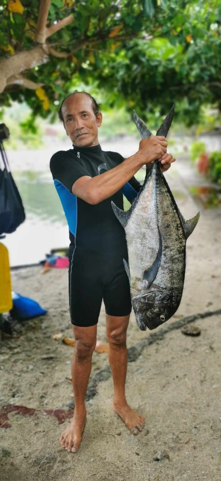 An indonesian man wearing a wetsuit stands on the beach holding a large fish up by the tail. In the background ocean, sand and equipment can be see laying under a large leafy tree.