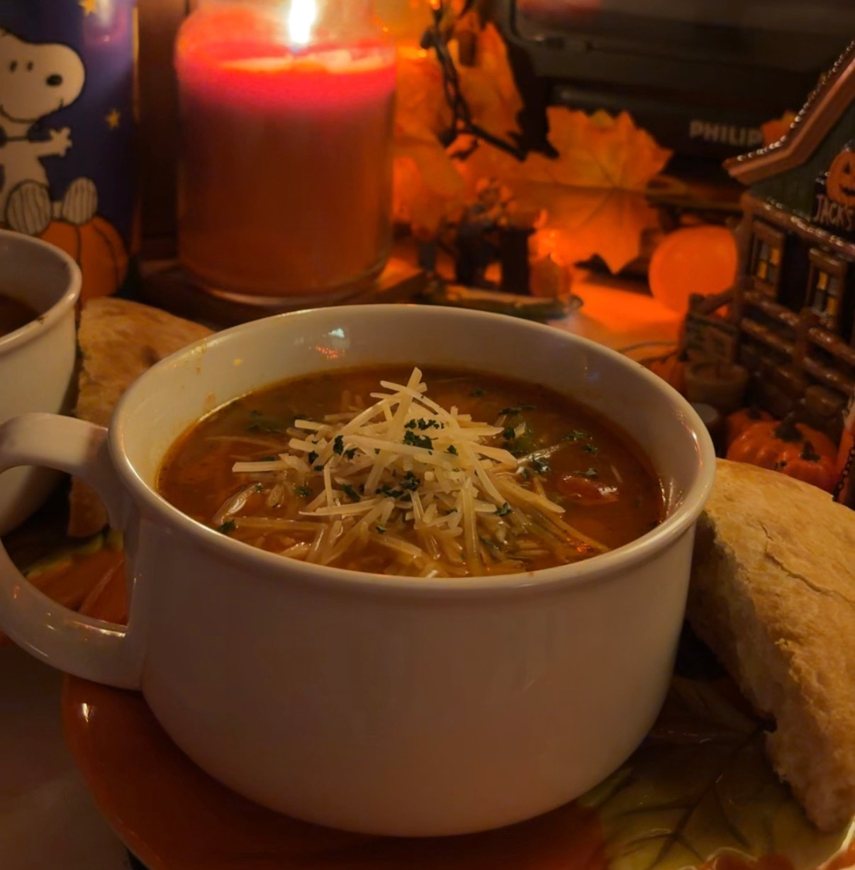 A cozy autumn scene with a warm bowl of soup topped with grated cheese and herbs, accompanied by a bread roll. The setting is illuminated by a glowing candle and surrounded by fall decorations, creating a perfect ambiance for a chilly evening.