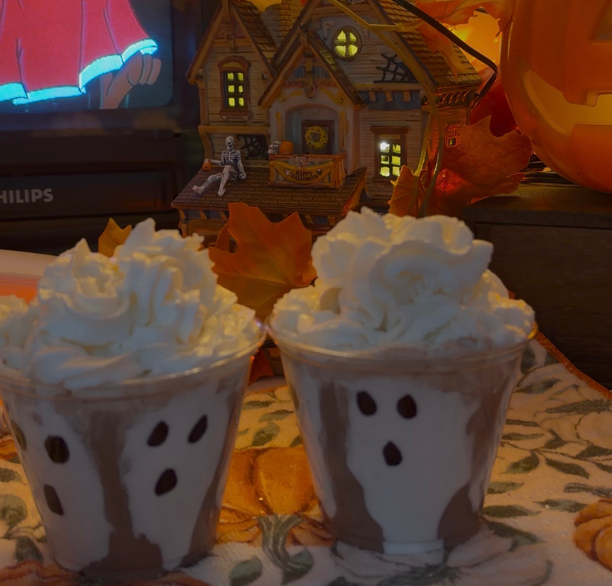 Cozy Halloween vibes with ghostly whipped cream topped drinks, surrounded by festive decorations and an old TV showing an animated classic.