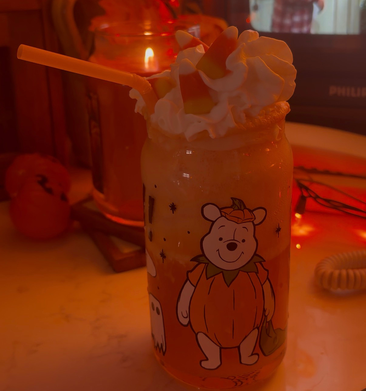Cozy autumn vibes with a spookily sweet beverage adorned with whipped cream and candy corn, a pumpkin-themed candle flickering nearby, and a classic movie setting the scene in warm, festive hues.