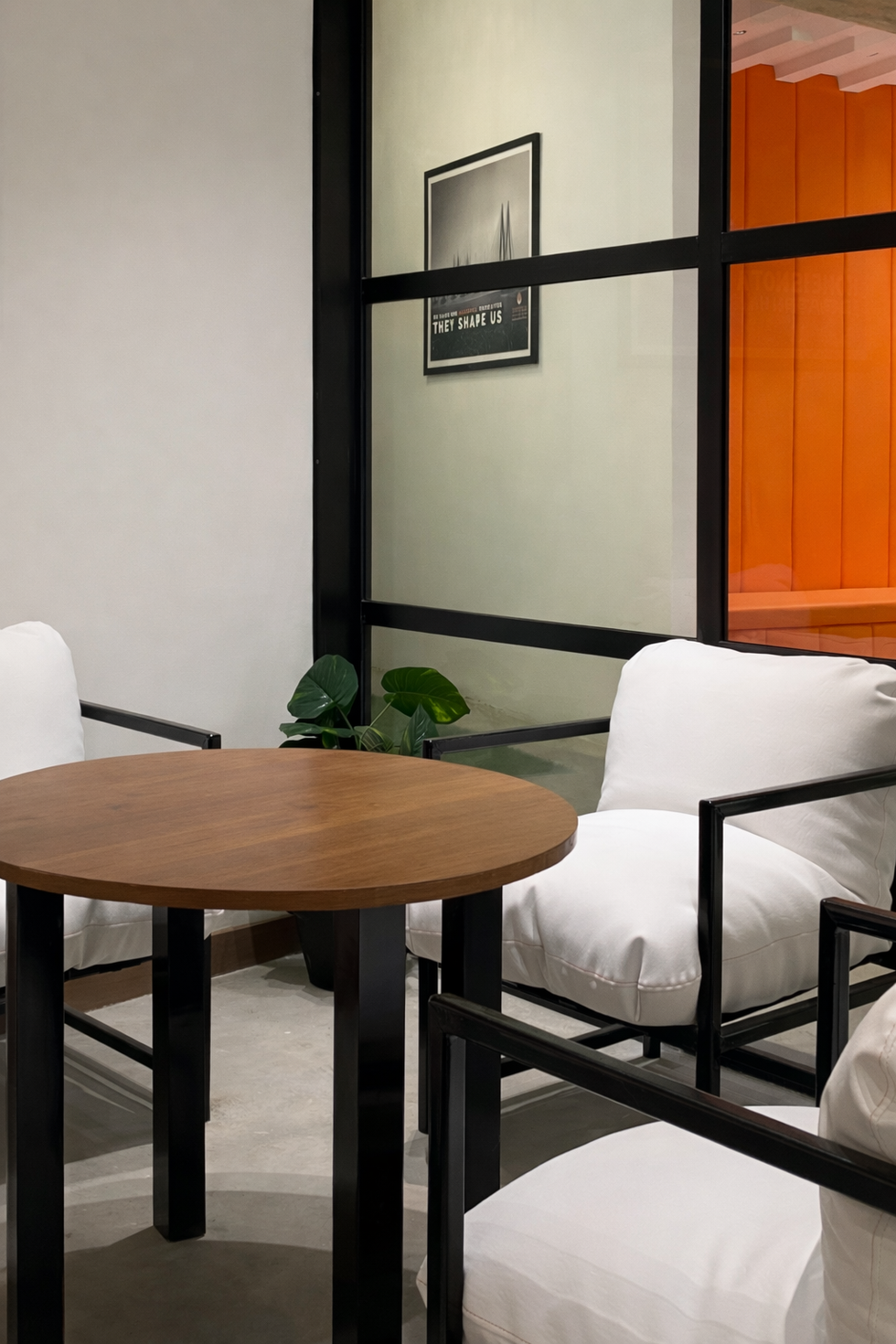 Modern sales lounge meeting area with round wooden table, white lounge chairs, glass partition, and orange accent seating designed by Woods & Bricks for Shreeji Builders