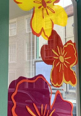 Close-up of large hand-painted flowers in yellow-green, orange and burgundy on a shop window, with a street and buildings reflected in the glass.