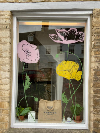Close-up of a shop window hand-painted with pink and yellow poppies, with potted plants and a branded tote bag on the sill inside.