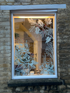 Shop window with a lit Christmas tree surrounded by hand-painted white poinsettia and holly illustrations, photographed at dusk.