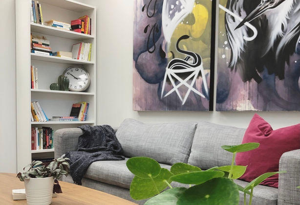 A photo showing the grey couch and large double panel painting of a pelican holding an umbrella. To the left of the couch is a white bookshelf filled with books, decorations and an analogue wall clock.