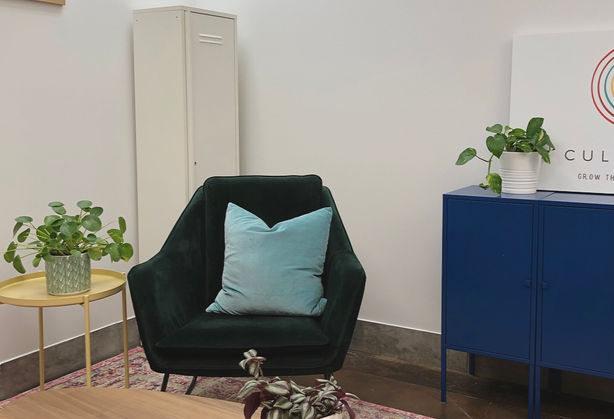 A photo of the other side of room one. It shows a dark green velvet chair with a teal pillow on it, a yellow side table, and a dark blue cabinet with a plant on it. In the foreground is a wood midcentury coffee table.