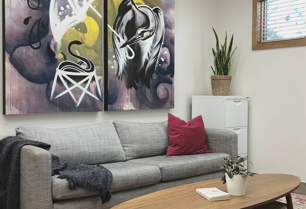 A photo of room one. It shows a large pink area rug, a midcentury wood coffee table, a grey couch and a double panel painting of a pelican holding an umbrella hung above the couch.
