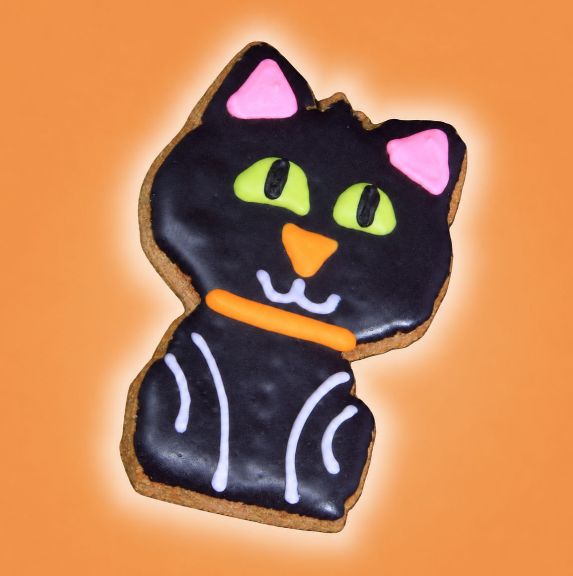 Black cat Halloween dog cookie with glossy black icing, green eyes and pink ears on warm orange background